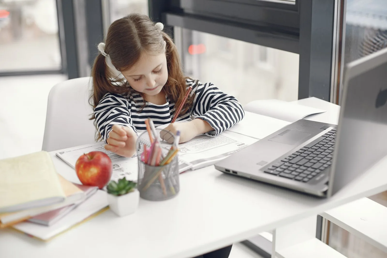 Niña escribe en su cuaderno en un escritorio ordenado, con un computador y una manzana