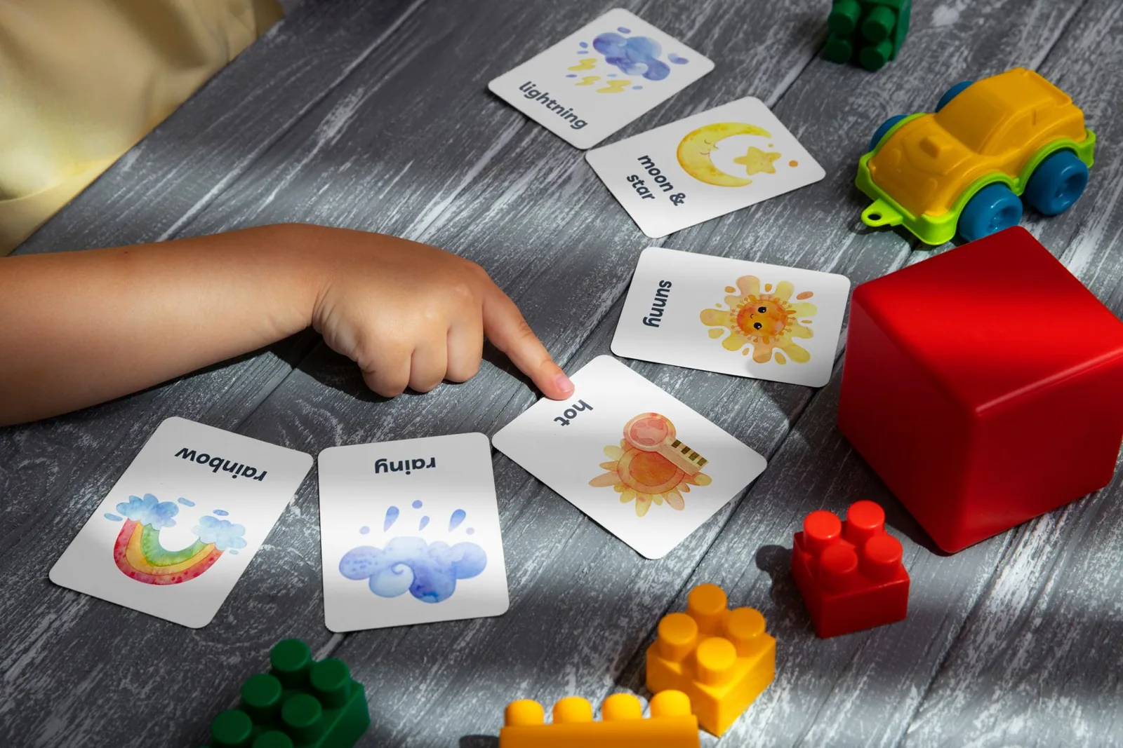 Tarjetas con palabras en inglés como rainbow y rainy sobre una mesa, junto a juguetes de colores