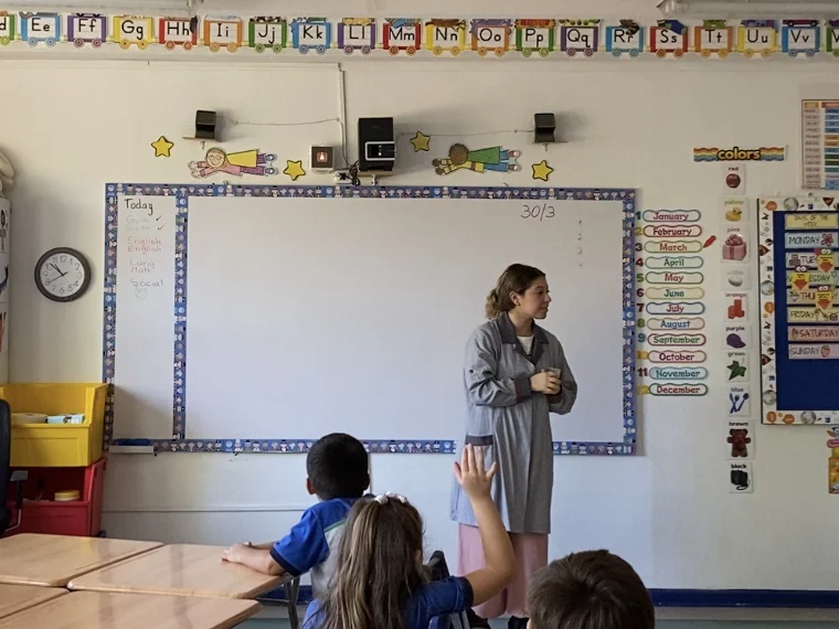Isidora Benito (Miss Isi) frente a la pizarra en una sala de básica bilingüe, con el abecedario en la pared y una alumna levantando la mano